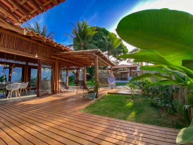 An expansive wooden deck is surrounded by lush greenery and tropical plants, leading to a serene pool area. Large glass doors enhance the connection between the indoor and outdoor spaces, while the thatched roof provides a natural aesthetic under the bright sky.