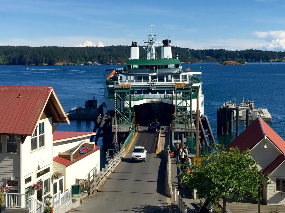 A ferry prepares to dock at the terminal, with vehicles lined up on the ramp. Surrounding buildings feature a mix of red roofs and light-colored exteriors. The calm water reflects the clear blue sky, framed by distant green islands.