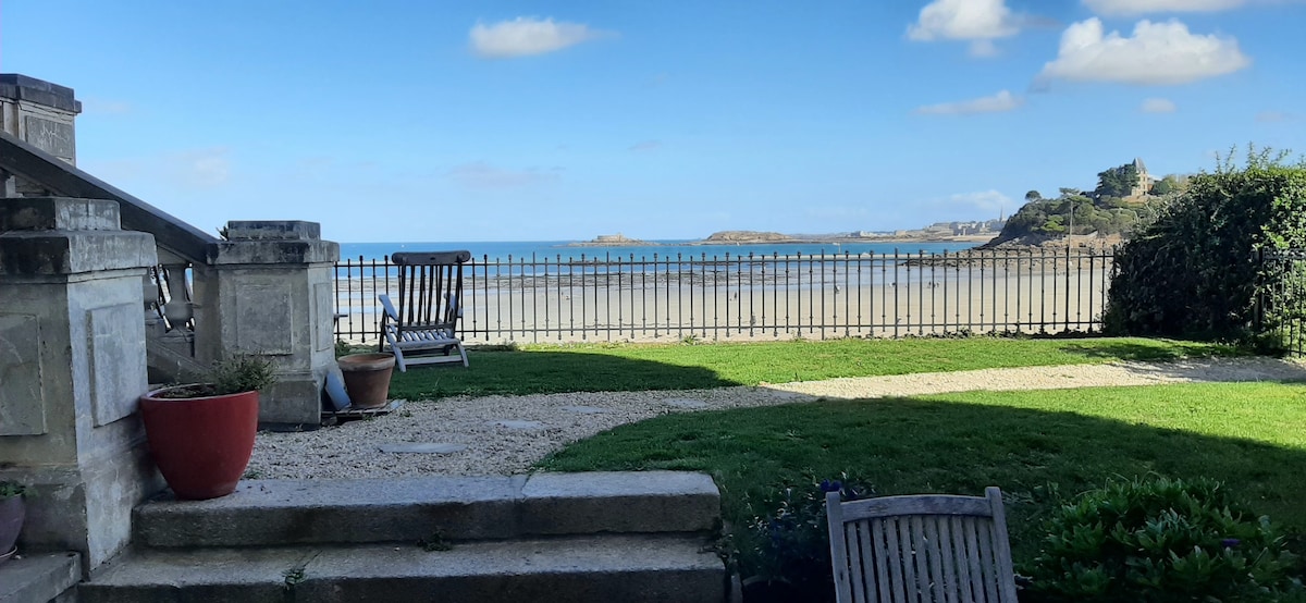 A serene outdoor area features a well-maintained garden with a view of the beach and ocean. Several lounge chairs are positioned on the grass, alongside decorative pots. The stone steps lead down from the property towards the beach, framed by a subtle fence.
