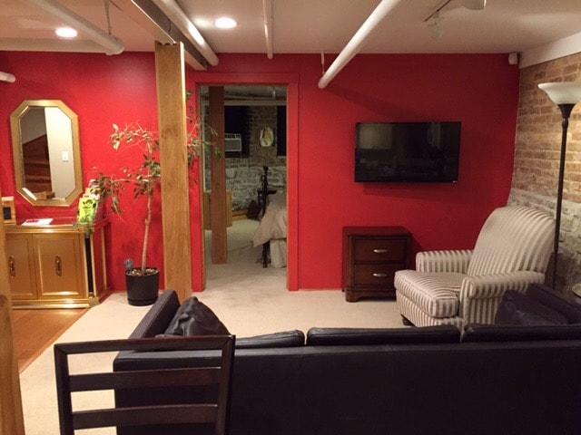 A cozy living area is highlighted by a red accent wall and brick details. A comfortable blue sofa faces a mounted television, with a striped chair nearby. A mirror and decorative plant are positioned against a stylish cabinet, complementing the space's inviting layout.