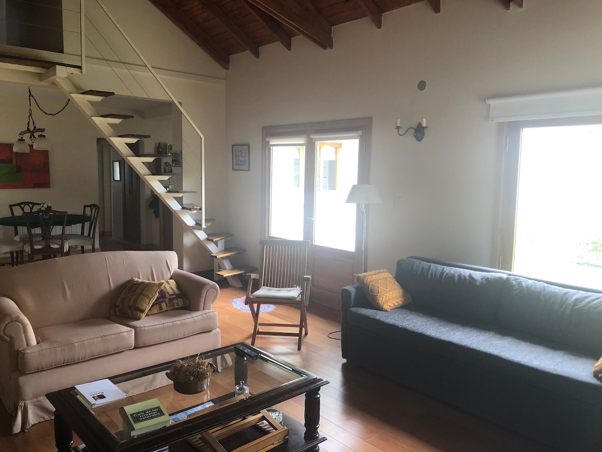A spacious living area features two sofas, a glass coffee table, and a wooden chair. Large windows allow natural light to fill the space, while wooden beams enhance the ceiling's height. A staircase is visible in the background, leading to an upper level.