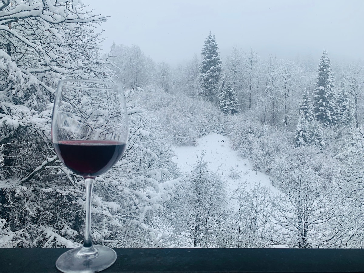 A glass of red wine is positioned on a snow-covered balcony railing, overlooking a serene winter landscape. Snow-laden trees fill the view, creating a tranquil atmosphere. The soft, muted tones of white and gray dominate the scene, evoking a sense of calmness.