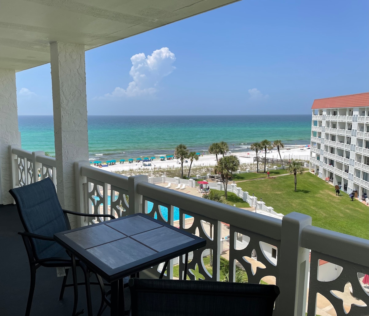 A spacious balcony is shown overlooking the Gulf of Mexico, showcasing vibrant turquoise waters and soft white sand. Two black chairs and a square table are positioned for relaxing moments, with a view of the pool area and palm trees in the foreground.