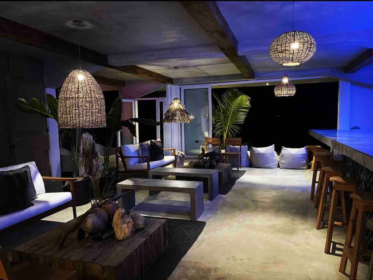 An outdoor lounge area is presented with stylish seating arrangements, including a mix of sofas and wooden chairs. Ambient lighting is provided by woven pendant lamps. Tropical plants and decorative elements enhance the environment, while a long bar counter adds functional space for dining or socializing.