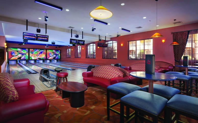 An indoor bowling alley is shown, featuring multiple lanes and vibrant artwork on the walls. Comfortable seating areas with red couches and round tables are positioned around, providing a relaxed atmosphere for guests to enjoy.