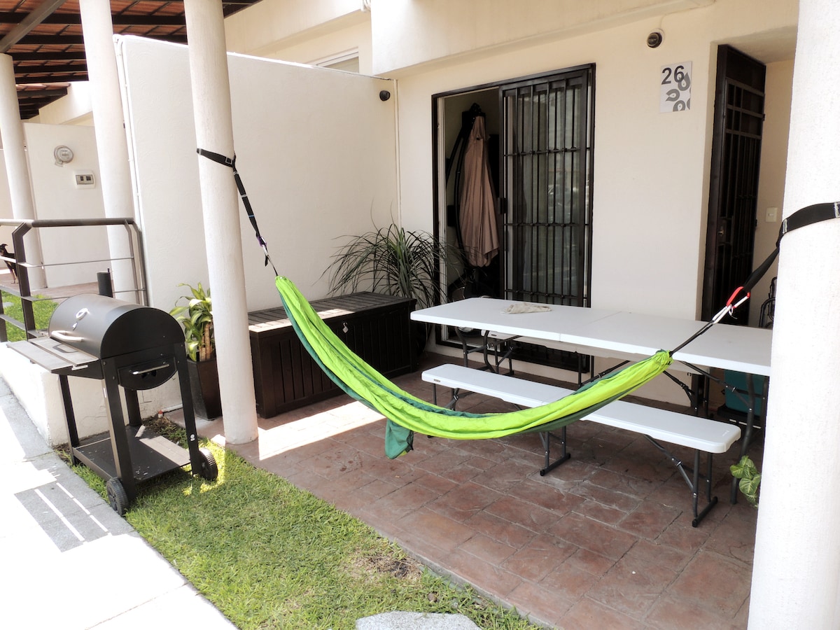A patio area features a vibrant green hammock suspended between two columns. A charcoal grill is positioned nearby on a rolling cart. A large, dark storage box sits against the wall, while a long, white table is set for gatherings, surrounded by spacious grass.