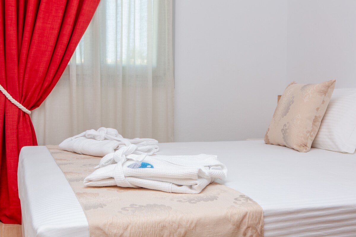 A clean sleeping area is displayed with a spacious bed featuring soft white linens. Coordinated bathrobes are neatly arranged on the bed. A bright red curtain complements the light coming from a window draped with sheer fabric.
