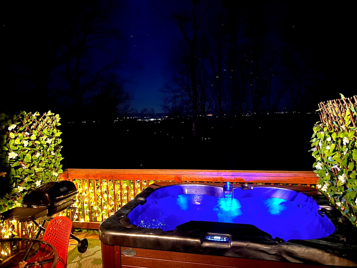 A hot tub offers a calming blue glow under the night sky, surrounded by lush greenery and twinkling lights. A grill is positioned nearby, and the distant city lights create a serene backdrop for evening relaxation.