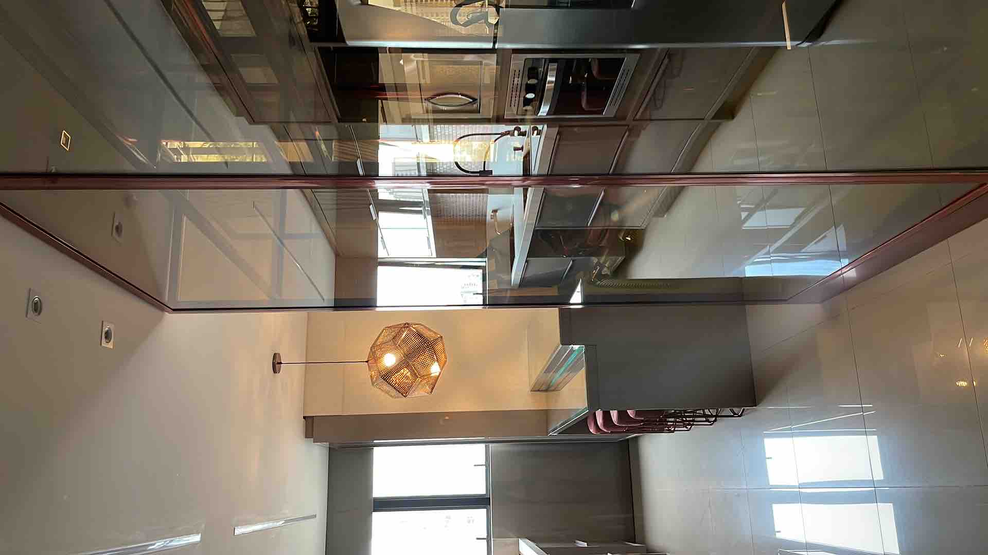 A modern kitchen features sleek cabinetry and stainless steel appliances. Expansive windows allow natural light to illuminate the space, reflecting off polished surfaces. An elegant pendant light hangs above a kitchen island, accompanied by several contemporary bar stools. The overall layout promotes an open and airy atmosphere.