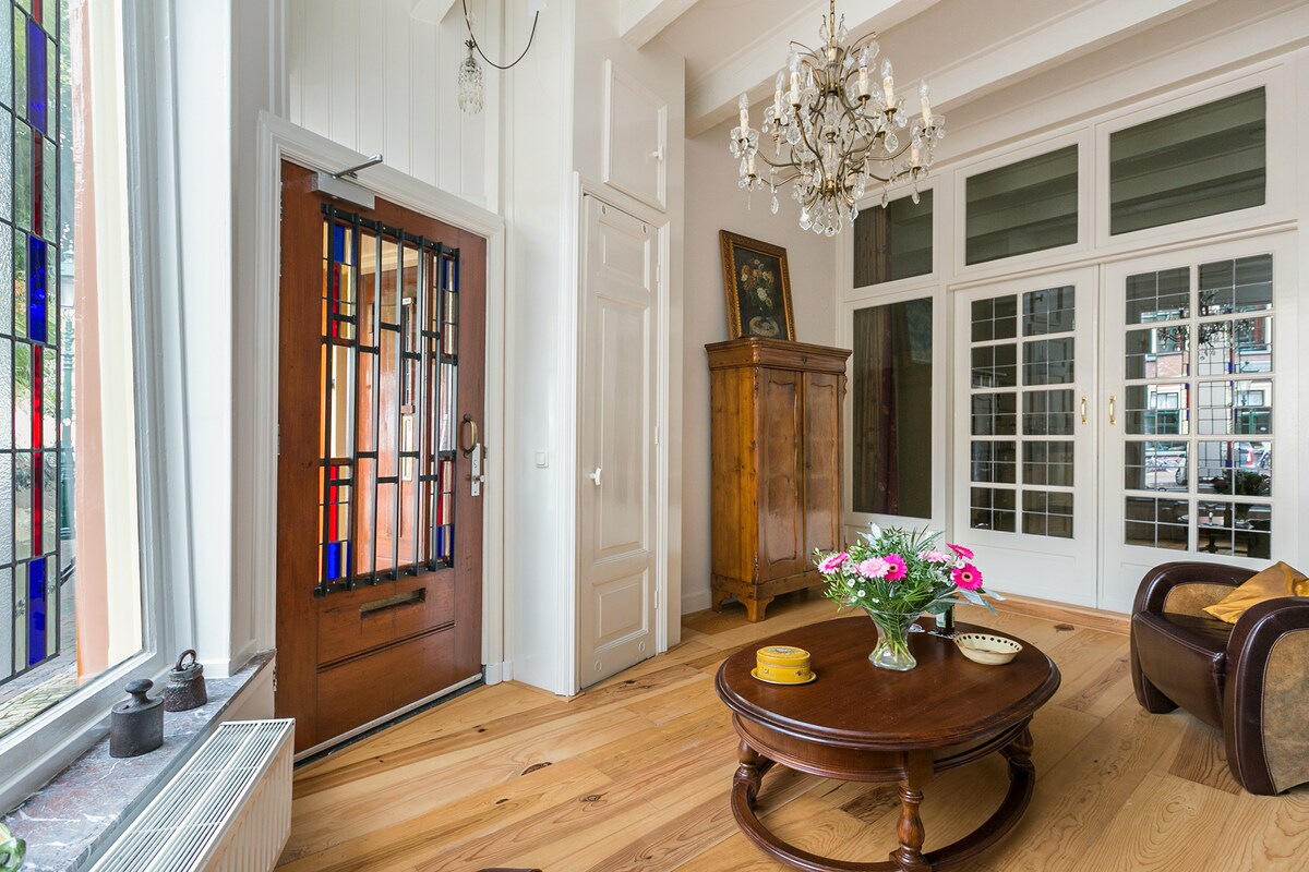 An elegant entryway features a stained glass door adorned with colorful designs. Inside, a round wooden coffee table sits centrally on a light wooden floor, accompanied by decorative flowers. A vintage wooden cabinet and a stylish armchair add character to the spacious area, illuminated by natural light.