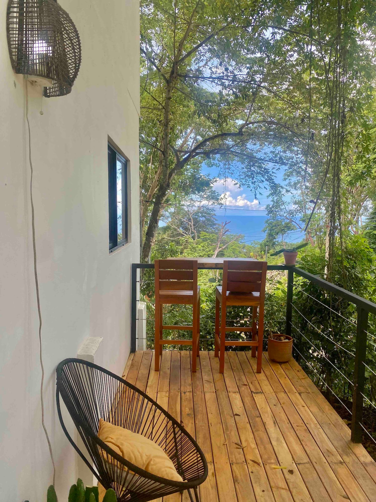 An outdoor deck made of wooden planks features two elevated bar stools beside a small potted plant. The surrounding greenery frames a view of the ocean in the distance, while sunlight filters through the trees, creating a serene atmosphere.