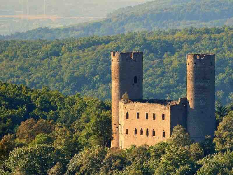 An ancient castle is nestled among lush green trees, showcasing its impressive stone walls and two prominent towers. The natural landscape extends into the distance, with rolling hills that blend seamlessly into the horizon.