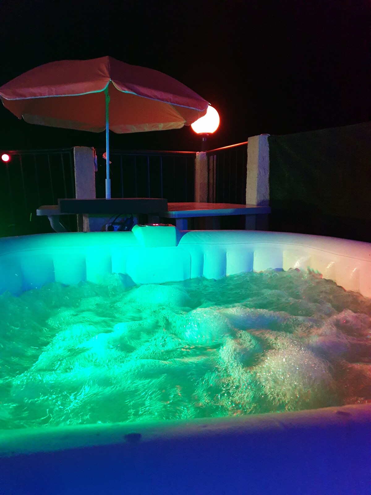 A jacuzzi is positioned on a terrace, illuminated by soft, colored lights. An umbrella provides shade above a small table situated nearby. The bubbling water creates a serene atmosphere, inviting relaxation.