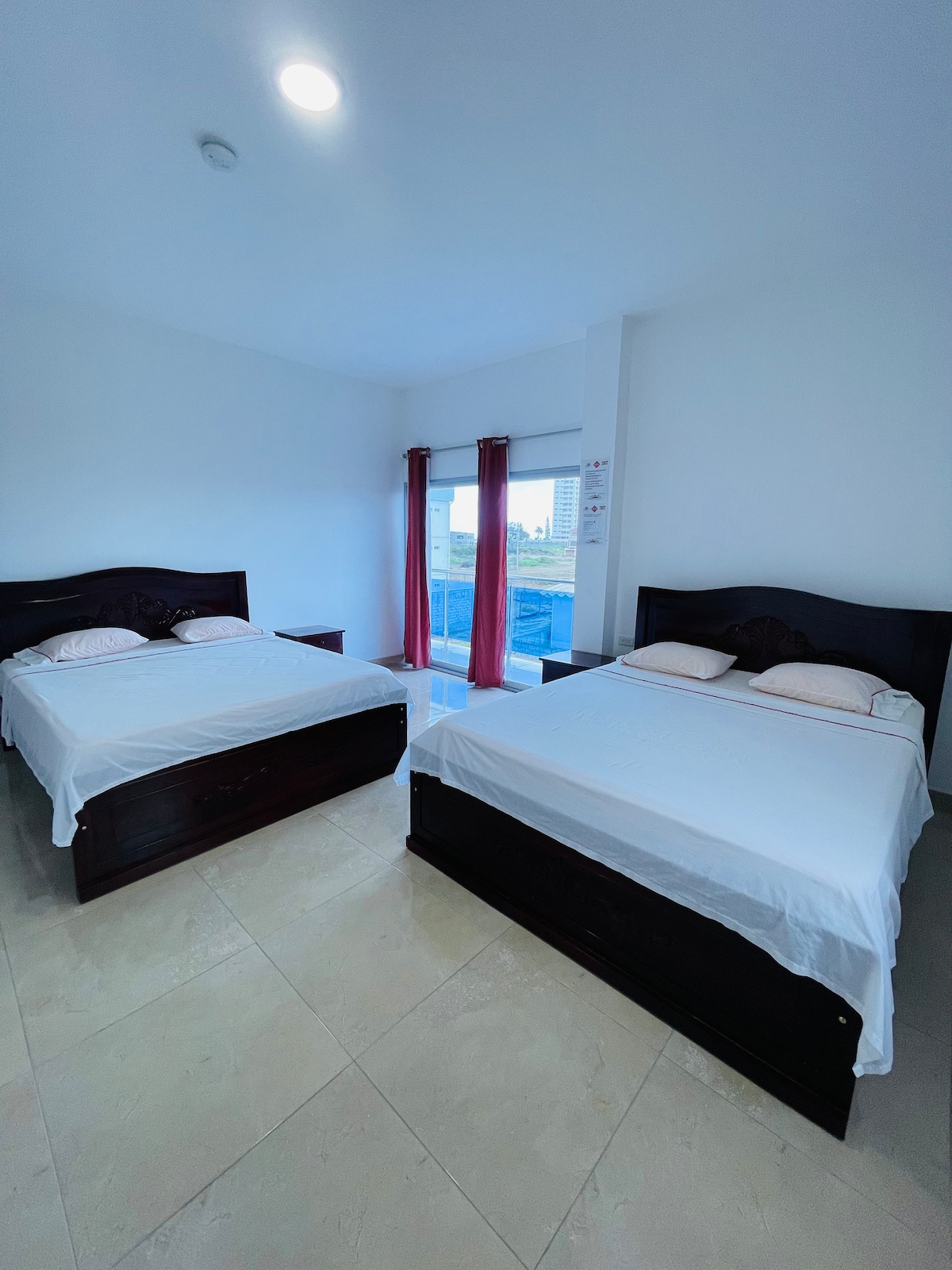 A bright, airy family room features two spacious beds with white linens, positioned against a wall. Natural light enters through a large window adorned with light red curtains, creating a calming atmosphere. The room is designed for relaxation and comfort.