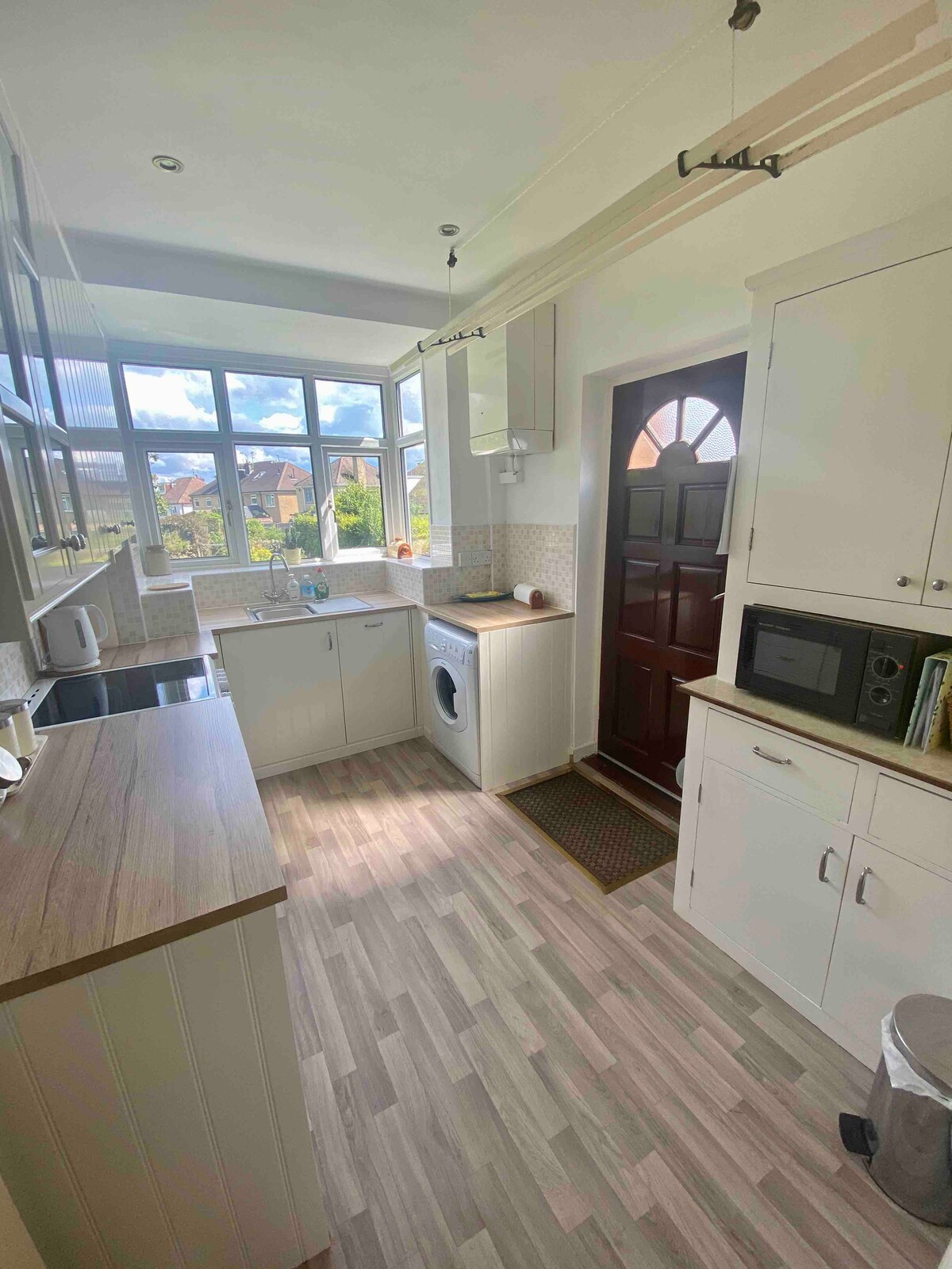 A bright, functional kitchen is displayed with light-colored cabinetry and a window that offers views of the outside. Essential appliances, including a washing machine and microwave, are positioned along the countertops, while natural light enhances the inviting atmosphere.