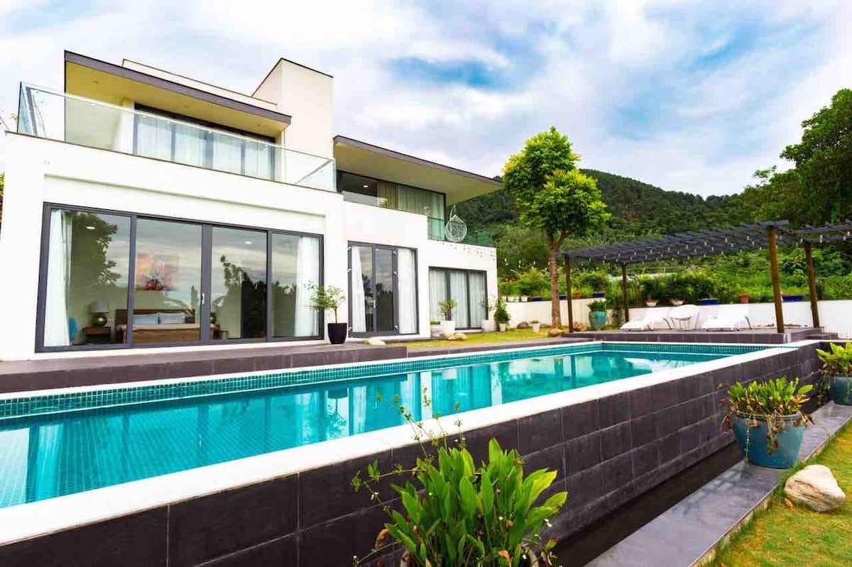 A modern house features large windows and a spacious balcony overlooking a serene pool. The outdoor area includes lush greenery and carefully arranged plants. A pergola nearby provides shade, enhancing the overall outdoor space.