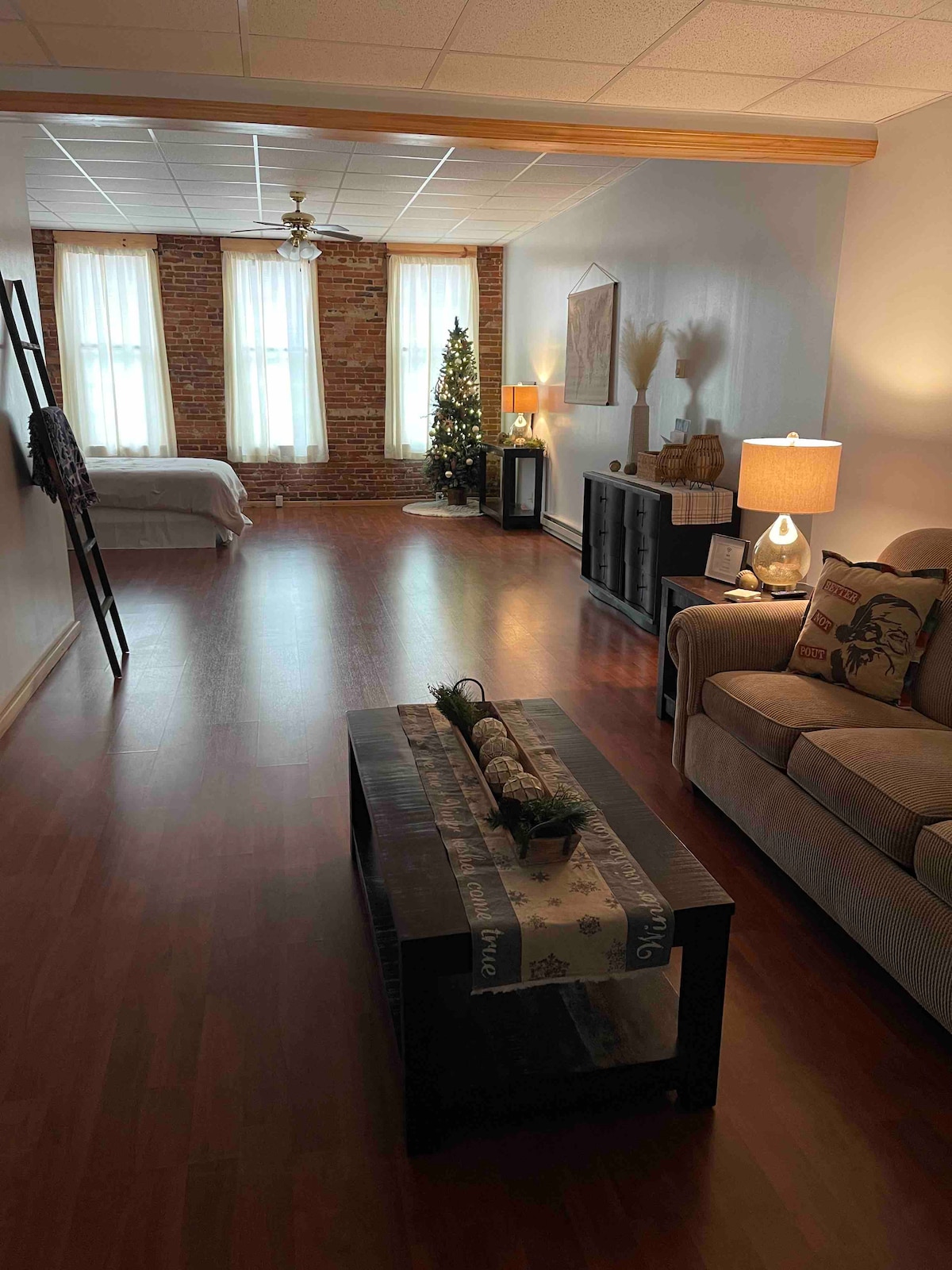 An open-concept loft space showcases a cozy seating area with a light-colored sofa and a coffee table adorned with decorative elements. Natural light filters through large windows, highlighting the exposed brick wall. A small evergreen tree adds a touch of seasonal charm.