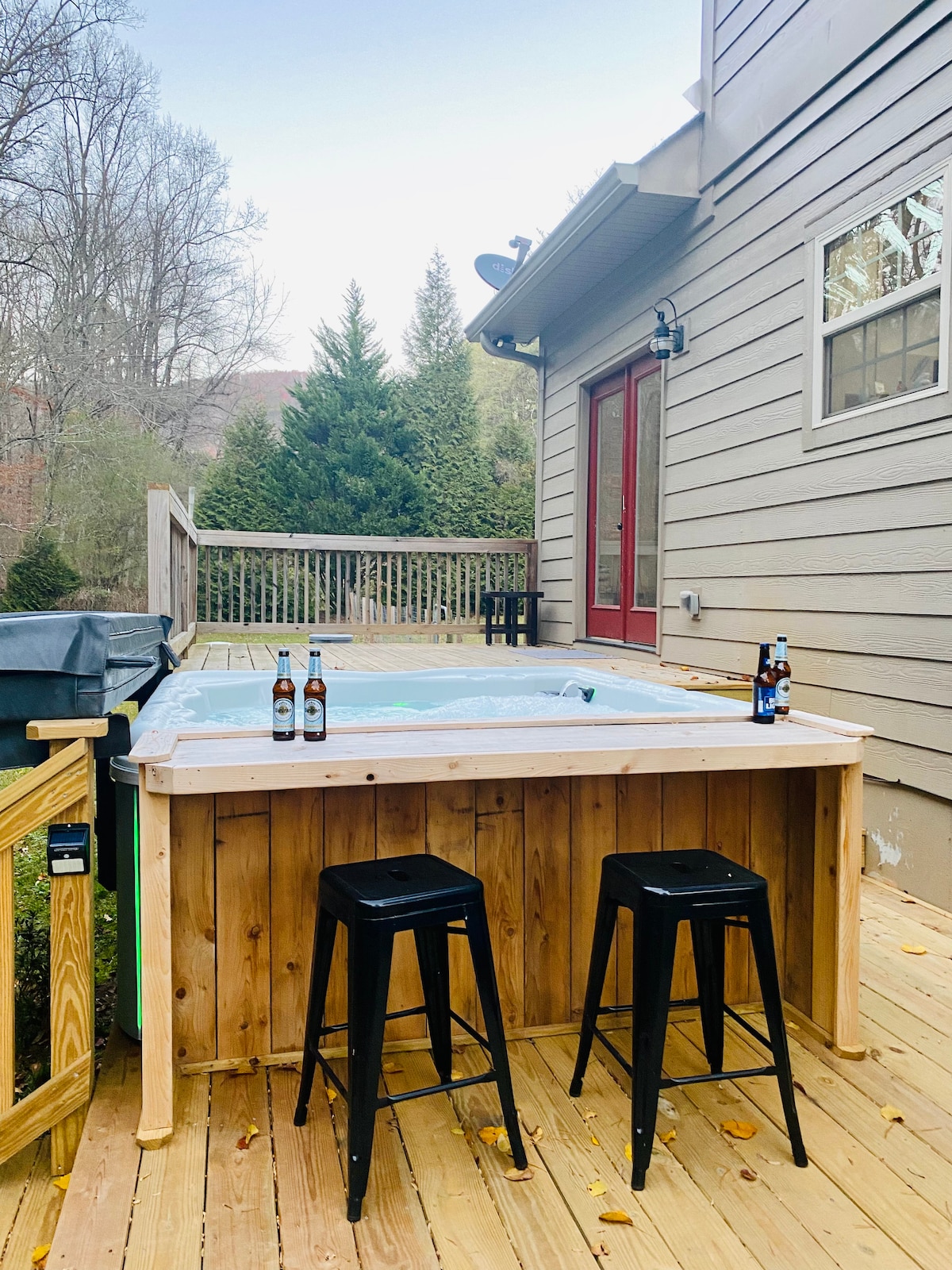 A wooden hot tub is situated on a spacious deck, accompanied by two black metal stools. Nearby, a BBQ grill is visible, and the area is surrounded by greenery, creating a serene outdoor space for relaxation.
