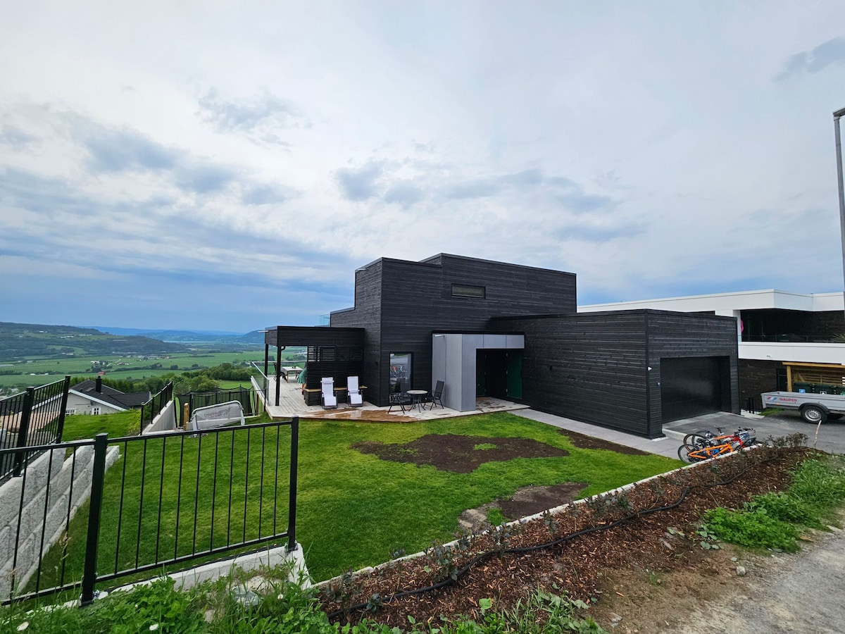 A modern black house is set against a backdrop of rolling hills, featuring a spacious lawn and a patio area with sun loungers. The structure showcases minimalist design elements and expansive windows, enhancing the outdoor views.