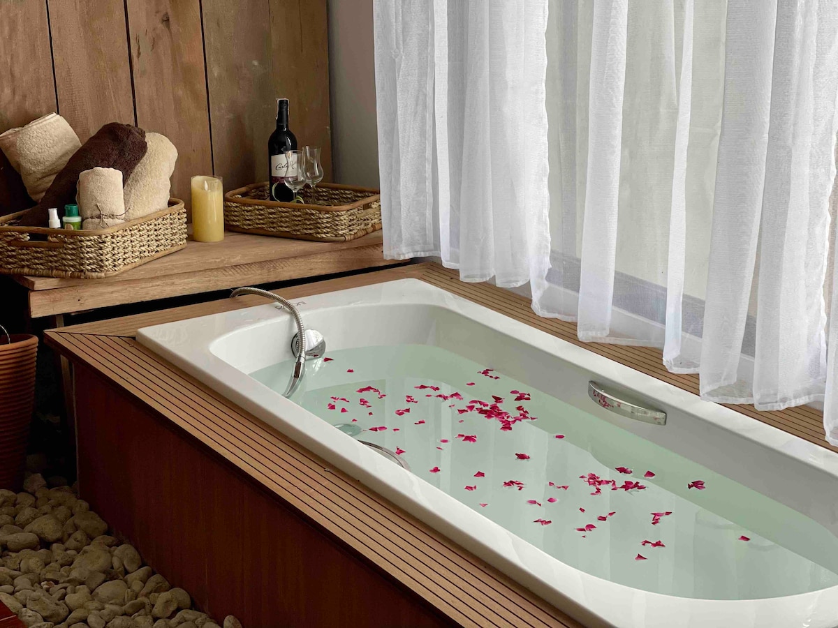 A well-appointed bathtub is surrounded by wooden paneling and pebbled flooring. Water is filled with scattered rose petals, creating a calming atmosphere. Soft, sheer curtains allow natural light to filter in, enhancing the serene ambiance of the bathing area.