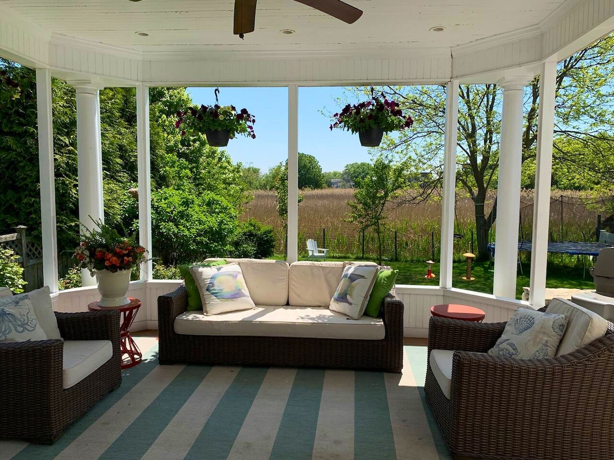 A spacious covered deck features comfortable seating with plush cushions, surrounded by large windows that offer views of the greenery outside. Potted plants hang from the ceiling, adding a touch of nature, while the striped rug enhances the inviting atmosphere.