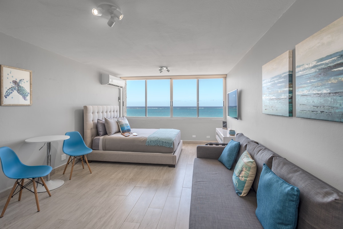 A spacious living area features a queen-sized bed and a comfortable full-sized sofa bed. Two blue chairs and a round table provide a cozy dining spot. Large windows offer an unobstructed ocean view, filling the space with natural light.