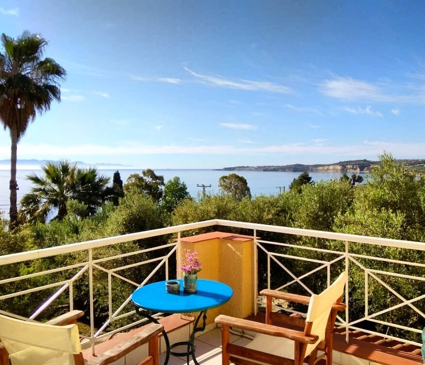A balcony view features a round blue table and two wooden chairs, surrounded by lush greenery. A small flower pot sits on the table, and a palm tree is visible to the left, framing the sweeping view of the sea and distant coastline.