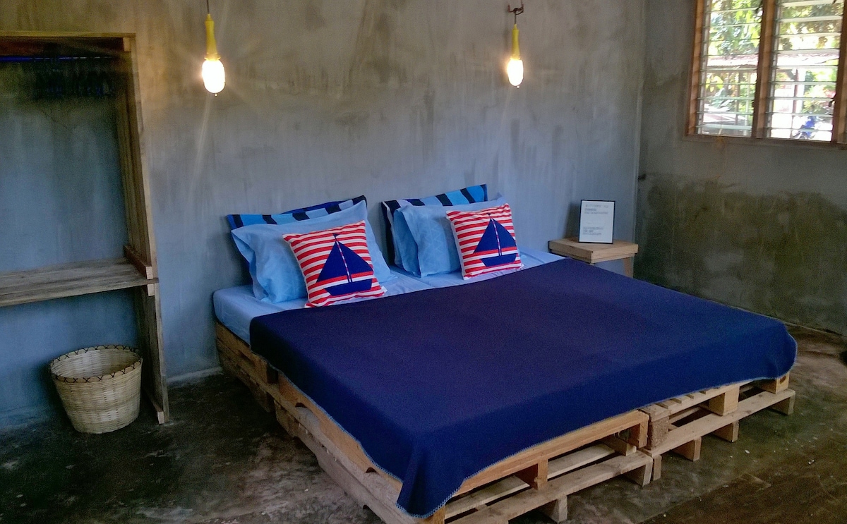 A spacious bed made from pallets is adorned with striped pillows and a dark blue blanket. The room features a concrete wall and a window with wooden slats, allowing natural light to enter. A woven basket is placed nearby for storage.