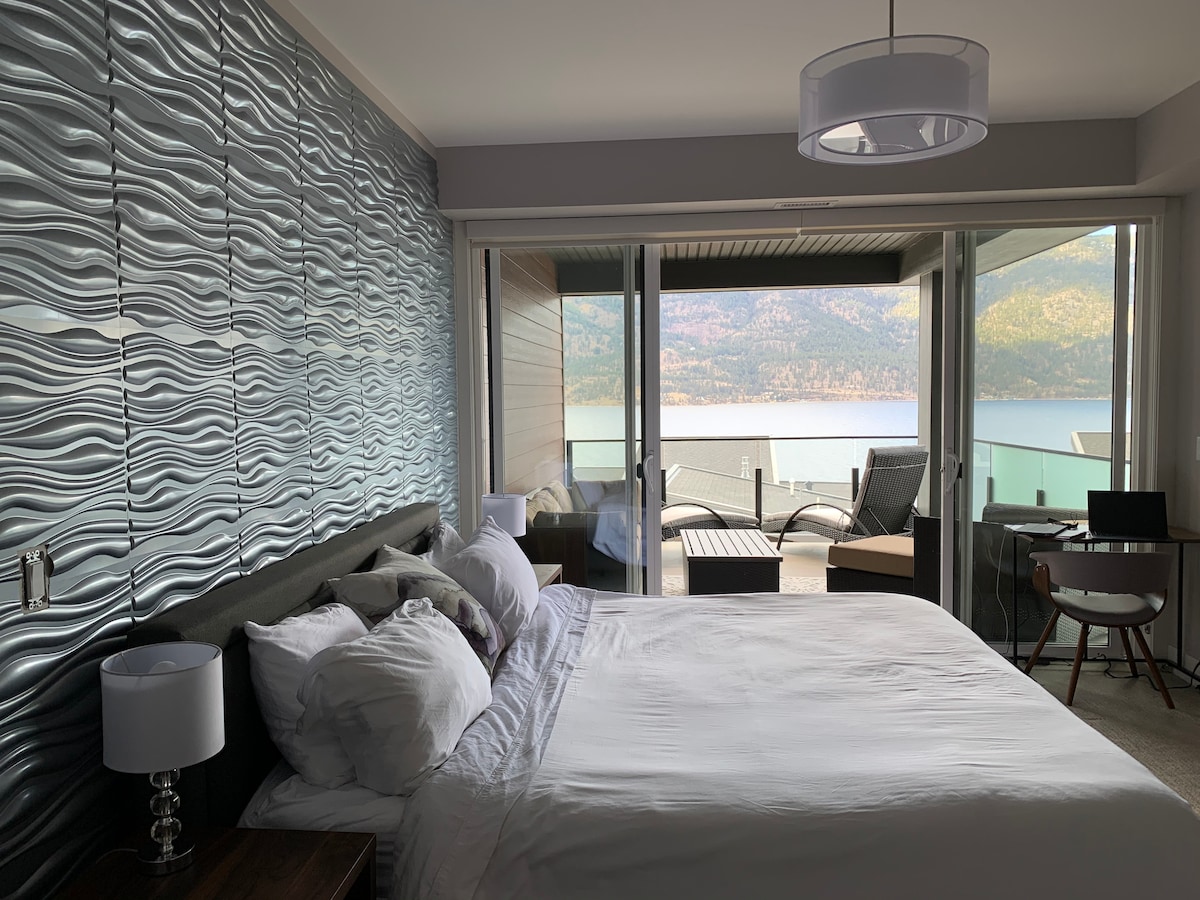 A spacious bedroom is showcased with large glass sliding doors leading to a private deck. The room features a king-sized bed with crisp white linens, accented by a textured wall in shades of blue. Comfortable lounge chairs are visible on the deck, offering lake views.