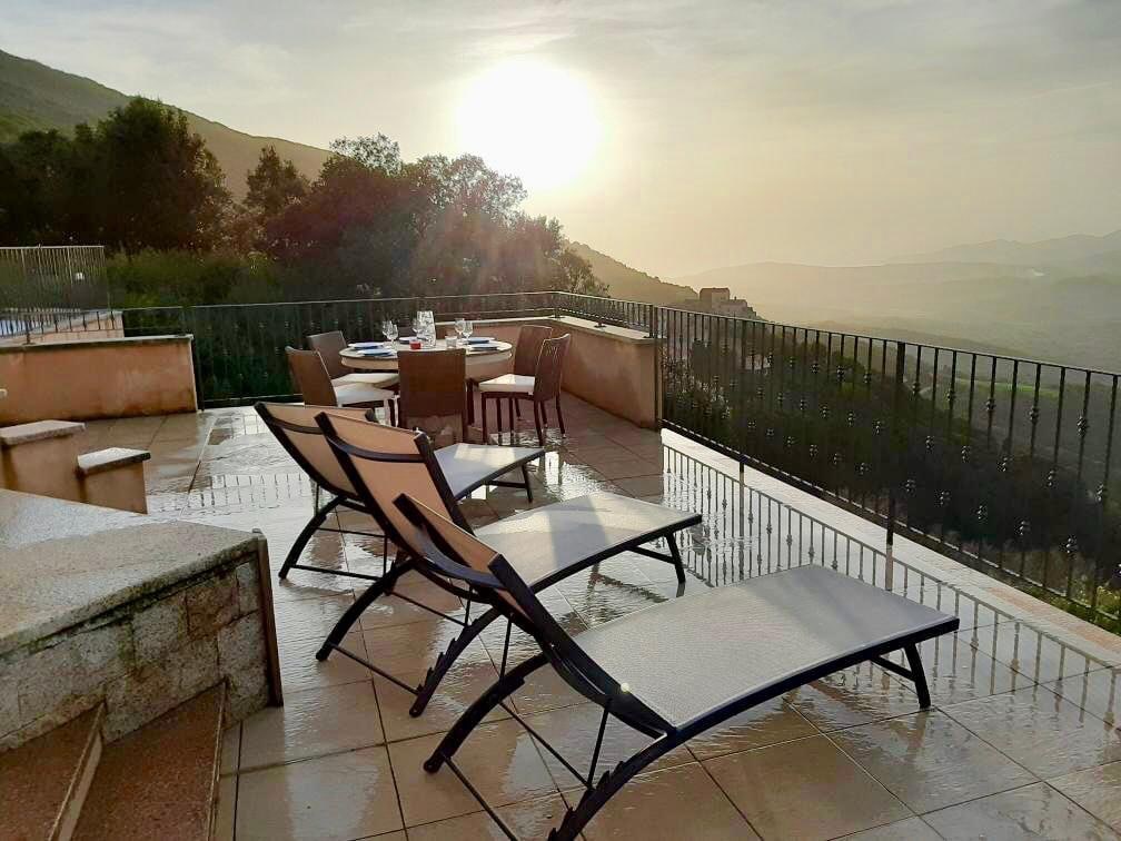 A spacious terrace features reclining loungers arranged for relaxation, with a dining table set nearby. The morning sun casts a warm glow over the landscape, enhancing the tranquil ambiance. The surrounding mountains provide a picturesque backdrop, while a railing ensures safety and unobstructed views.