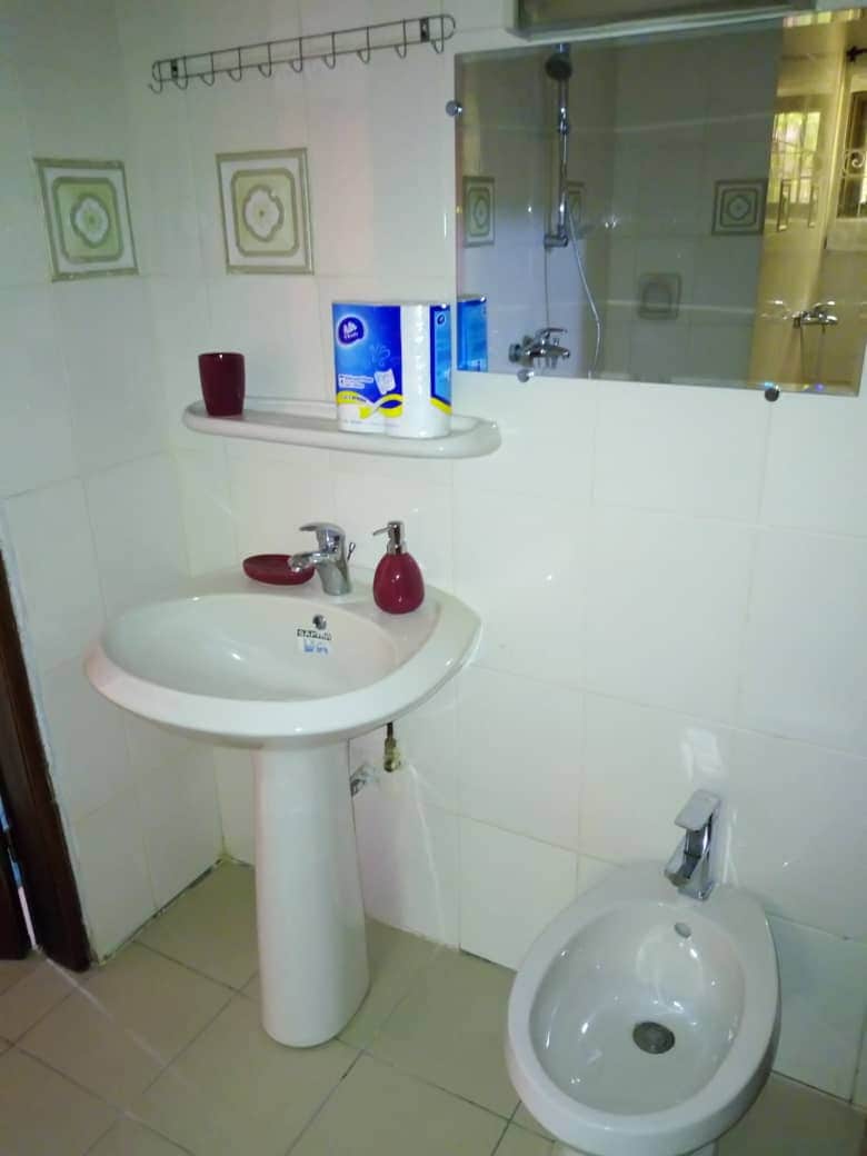 A bathroom is featured with a white pedestal sink and a bidet, both set against light-colored tiled walls. A mirror reflects the space, and various toiletries are neatly placed on the white shelf above the sink.