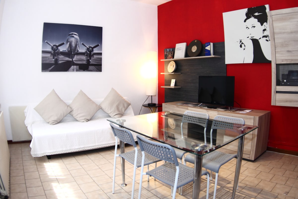 A living area is presented with a glass dining table surrounded by six grey chairs. A white sofa with large cushions is positioned across from a wooden entertainment unit featuring a television. Art pieces adorn the walls, one prominently displaying a vintage aircraft.