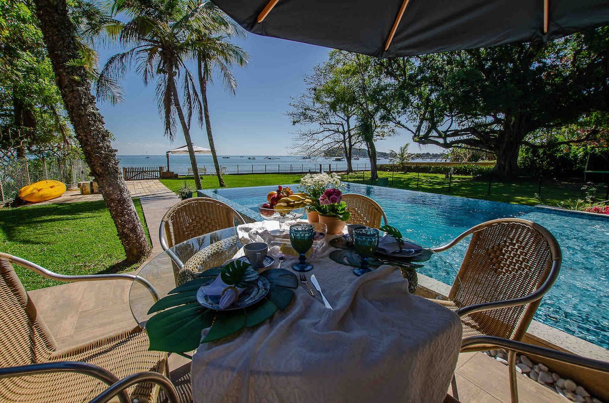 An outdoor dining area is set up with a table featuring elegant dinnerware and colorful flowers. Surrounding it are four woven chairs, with a view of a sparkling swimming pool and lush greenery. Palm trees frame the scene, while the ocean can be seen in the distance.