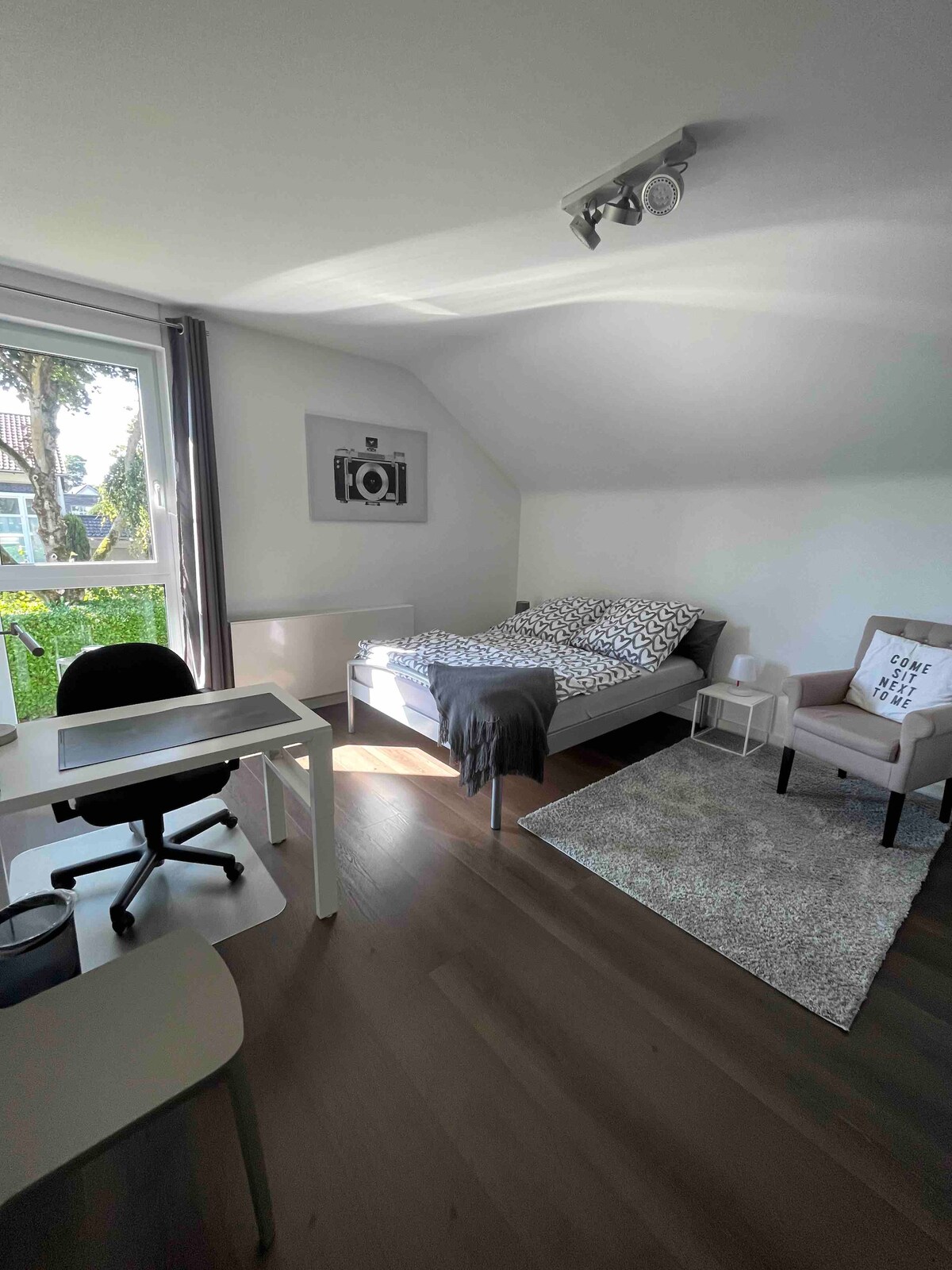 A bright sleeping area is featured, equipped with a spacious bed dressed in patterned bedding. Natural light enters through large windows, enhancing the room's open feel. A desk with an ergonomic chair, a comfortable armchair, and a soft area rug add to the inviting atmosphere.