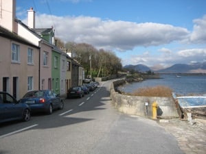 A quiet coastal road lined with pastel-colored houses runs alongside a shimmering body of water. Parked cars are seen on the left, and stone walls border the right side, offering views of distant mountains beneath a partly cloudy sky.