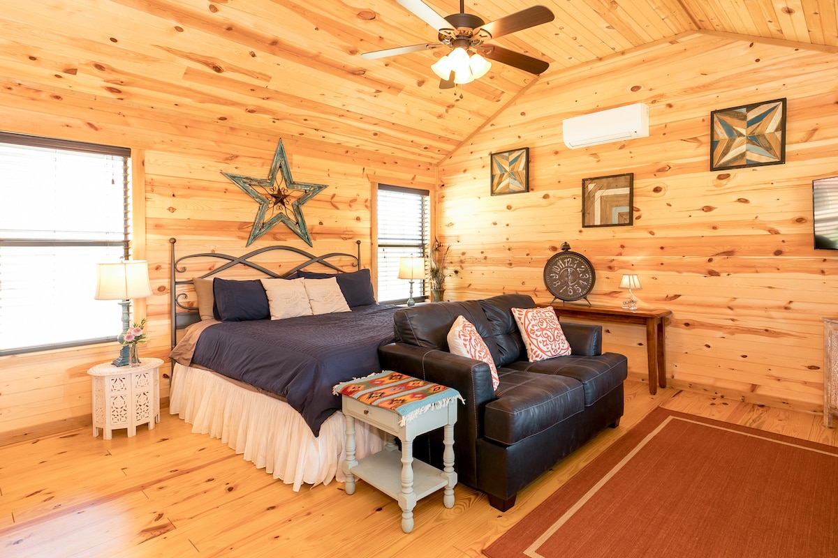 A warm wooden interior features a spacious king bed with dark bedding and a decorative star above. A charming sofa sits opposite a small table, with lamps providing soft lighting. Windows allow natural light to enhance the inviting atmosphere of the room.