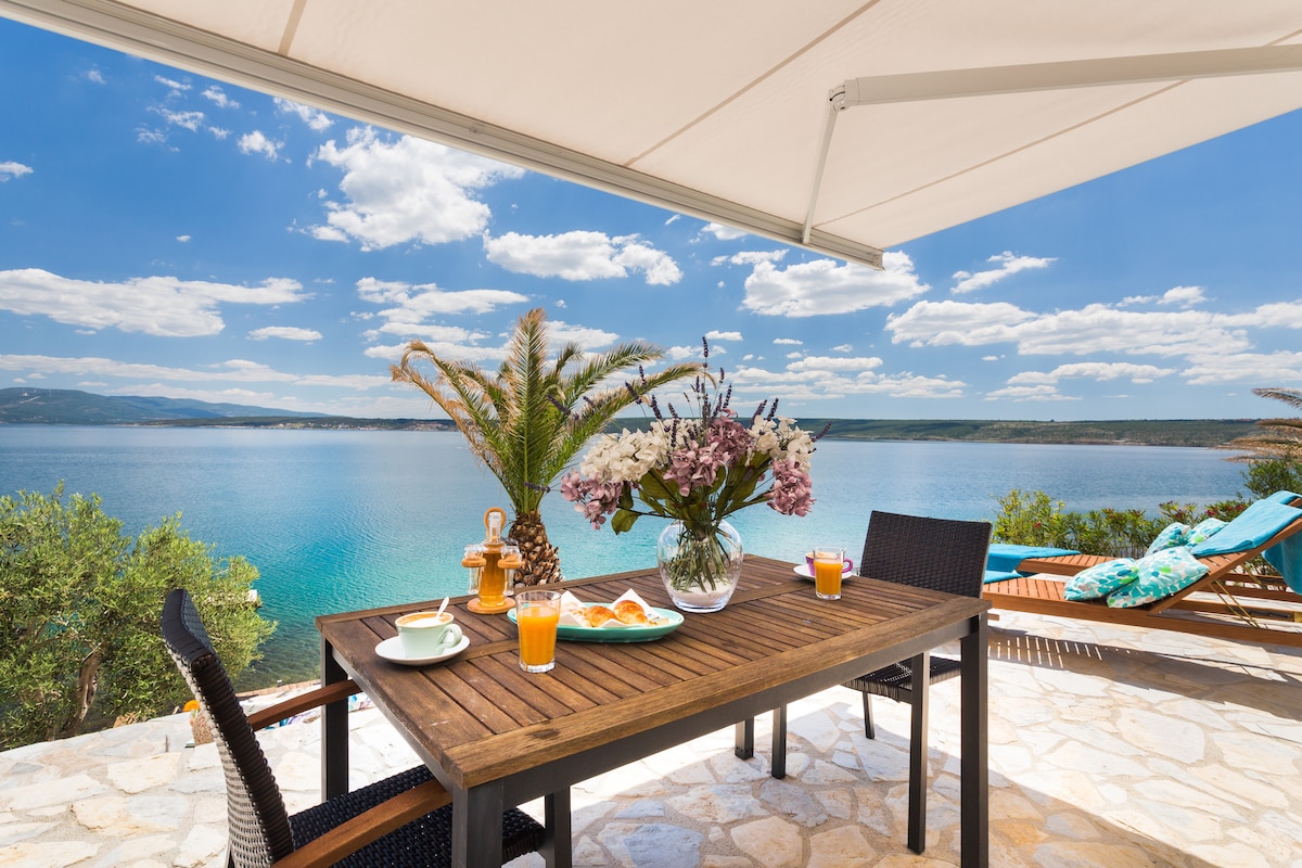 A wooden dining table set outside showcases a vase filled with fresh flowers. Two glasses of orange juice and a plate of pastries are arranged on the table. Sun loungers are positioned nearby, with a panoramic view of the calm water and distant hills under a partly cloudy sky.