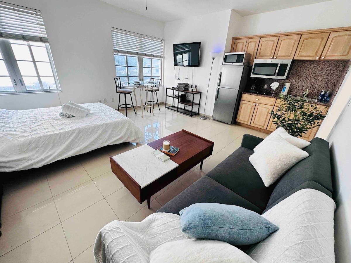 A spacious studio is presented with natural light filtering through large windows. The space includes a queen-sized bed with white bedding, a couch, and a coffee table. A fully equipped kitchen is visible, along with a dining table and a flat-screen TV mounted on the wall.