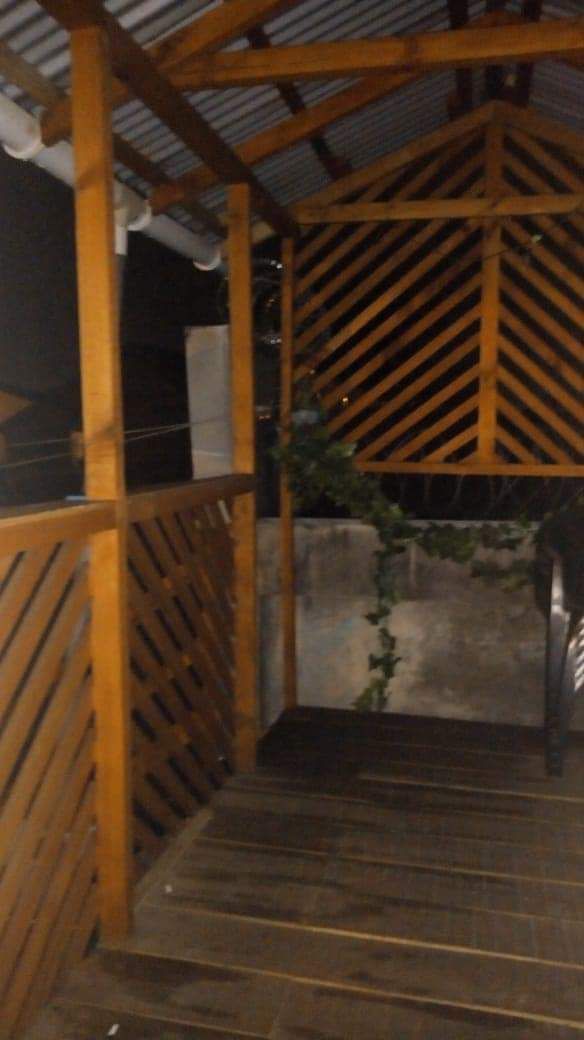 An elevated wooden terrace is shown, featuring a slatted design that enhances visual interest. The structure is surrounded by greenery, with a clear view of a wall at the end. Soft lighting suggests a relaxed evening ambiance.