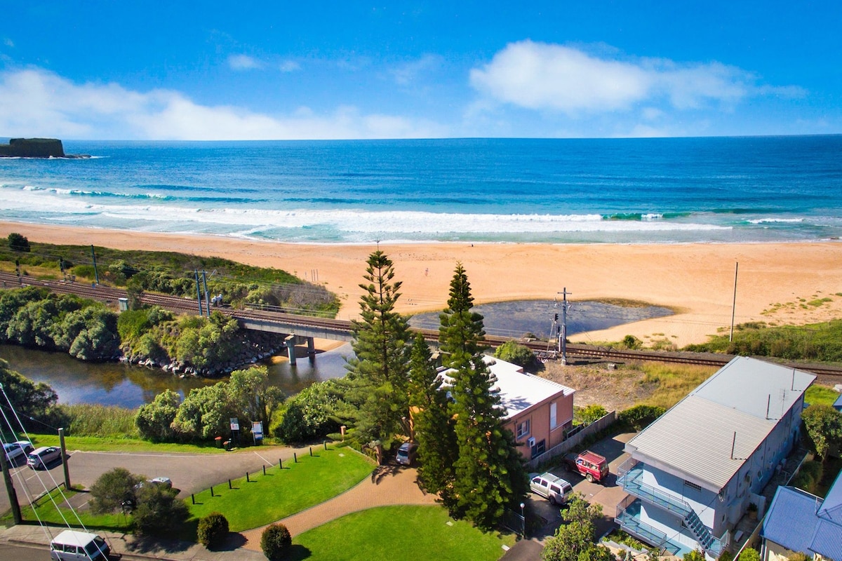 An expansive view of the sandy beach stretches across the foreground, meeting the tranquil ocean waves. Lush greenery lines the edge of the beach, while a railway runs parallel to the coast. The horizon features clear blue skies, enhancing the serene coastal atmosphere.