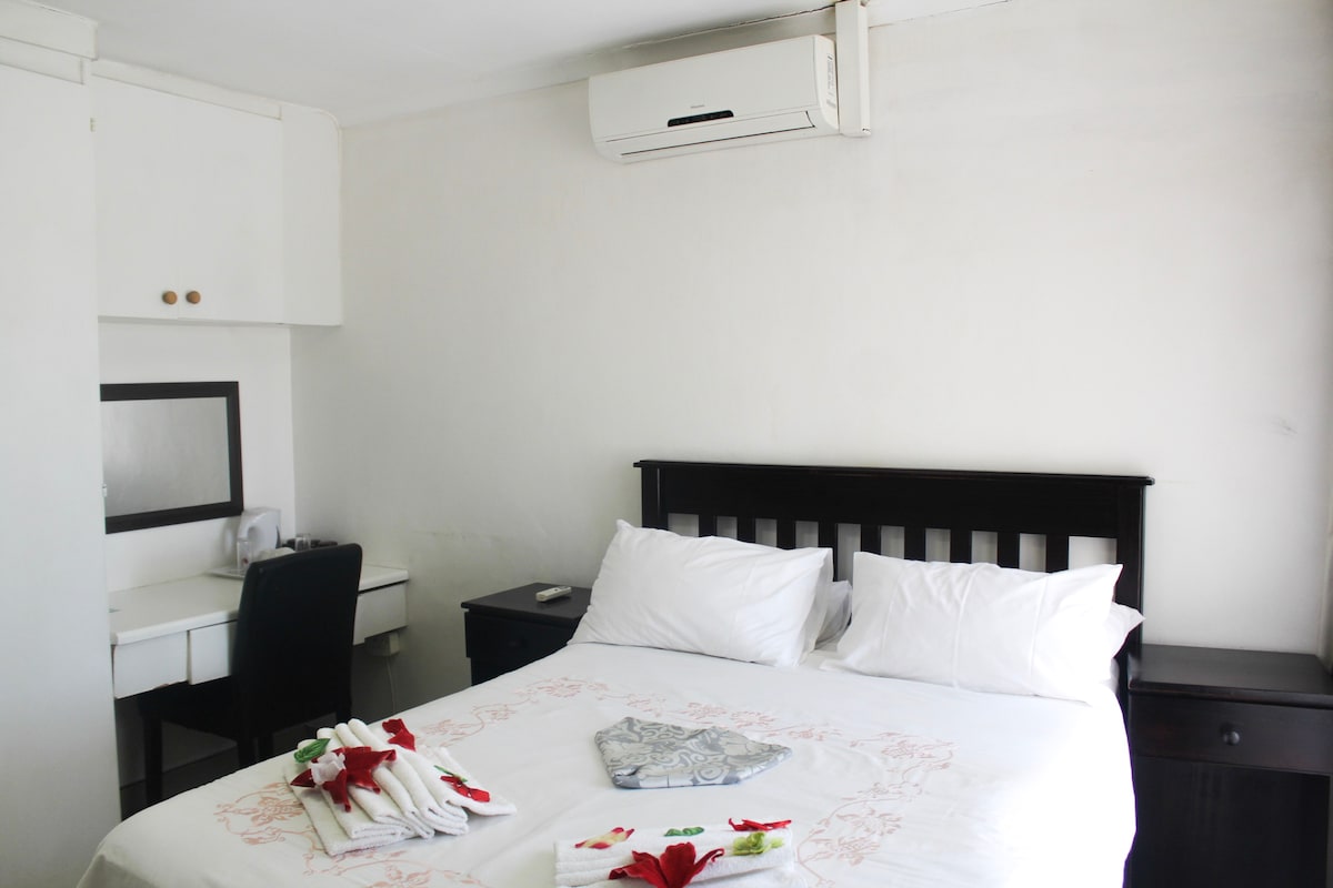 A simple and clean room features a spacious bed with white linens and decorative accents. A desk with a chair is positioned nearby, along with a mirror. Air conditioning is installed on the wall, contributing to the comfort of the room.