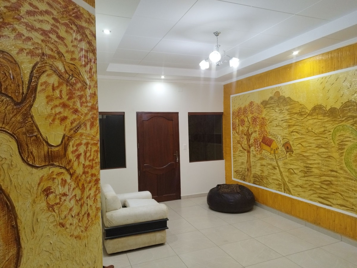 A spacious living area features decorative murals on the walls, showcasing natural scenes. A light-colored sofa is positioned near a large artistic panel. Warm lighting from overhead fixtures enhances the inviting ambiance of the room.