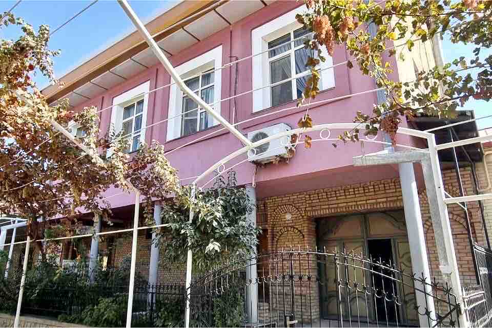 The exterior of a two-story building is shown, featuring a pink facade and white window frames. Lush greenery and grapevines are visible, complementing the welcoming entrance area. A decorative iron fence encircles the property, enhancing its charm.
