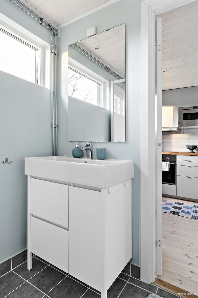 The modern bathroom features a sleek white sink unit with storage underneath. A large mirror is mounted above the sink, complemented by a light blue wall and windows allowing natural light. The floor is tiled in a dark pattern, enhancing the room's clean design.