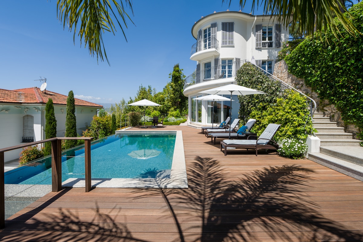 A large infinity pool is bordered by a wooden deck, featuring several sun loungers and umbrellas for shade. The villa's stylish architecture is complemented by lush greenery and flower beds, creating a serene outdoor environment. Clear blue skies enhance the overall tranquility.