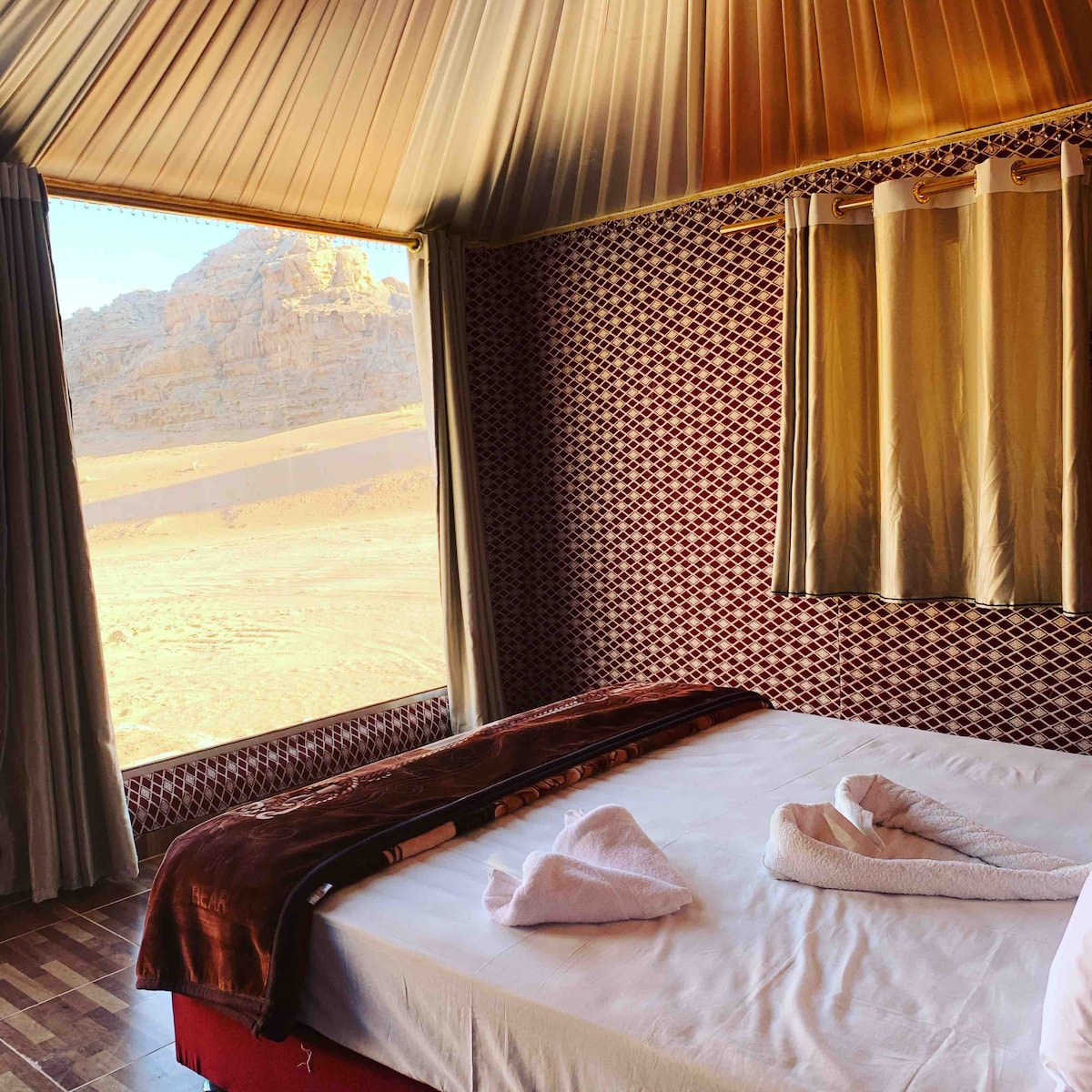 A spacious tent interior features a simple bed adorned with neatly arranged towels. Natural light floods in from a large window, offering a view of the surrounding desert landscape and rocky formations. Earthy tones and patterned walls create a serene atmosphere.