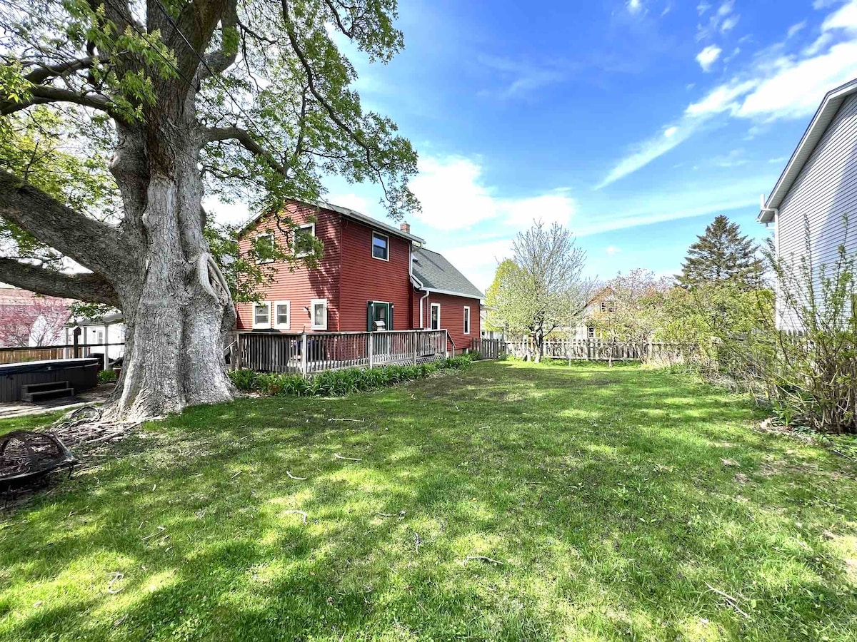 A spacious yard surrounds the historic Red Farmhouse, featuring well-maintained grass and mature trees. A wooden fence encloses the backyard, providing privacy. The clear sky and leafy branches create a serene setting, ideal for outdoor activities and relaxation.