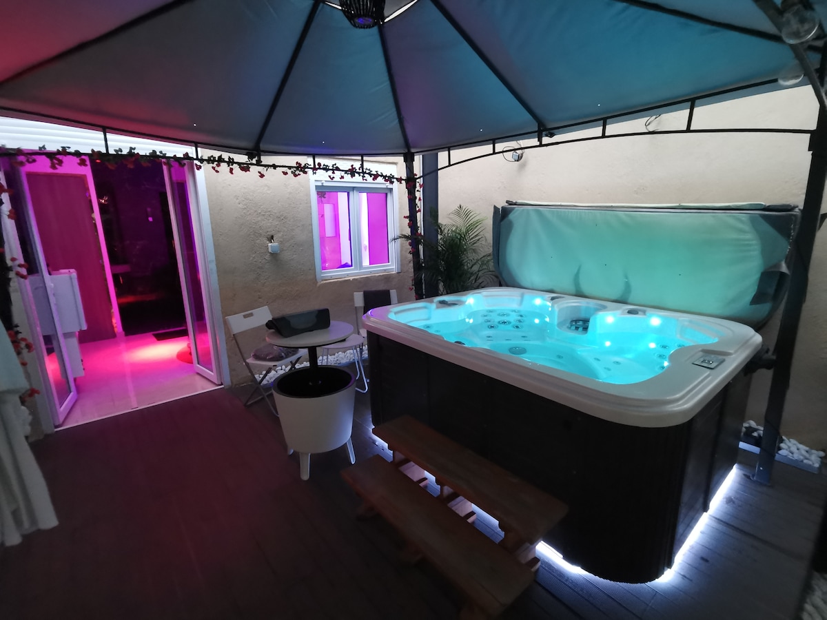 A spacious outdoor area features a hot tub surrounded by slatted decking. Soft lighting illuminates the space, complemented by a canopy above. Nearby, a small table and chairs are positioned, and sliding glass doors lead to an interior area, enhancing the cozy ambiance.