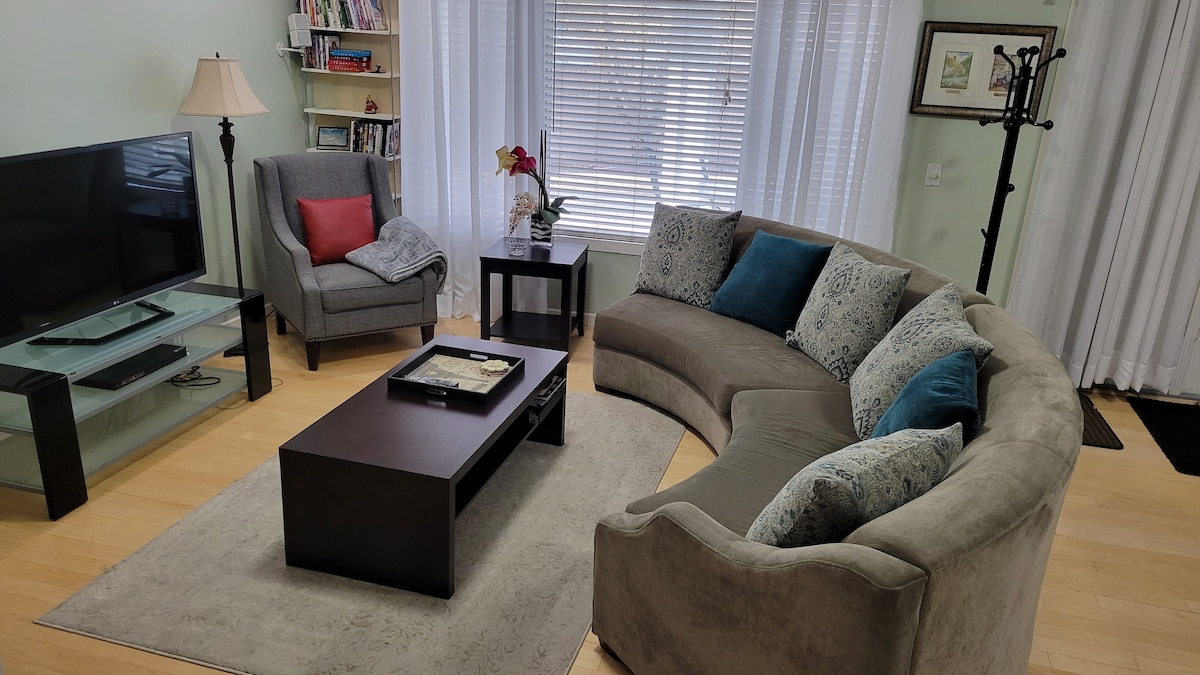 A spacious living area features a curved sectional sofa adorned with cushions and a coffee table at its center. A TV is positioned on a glass stand, complemented by a stylish armchair. Natural light filters through sheer curtains, enhancing the space's inviting appeal.