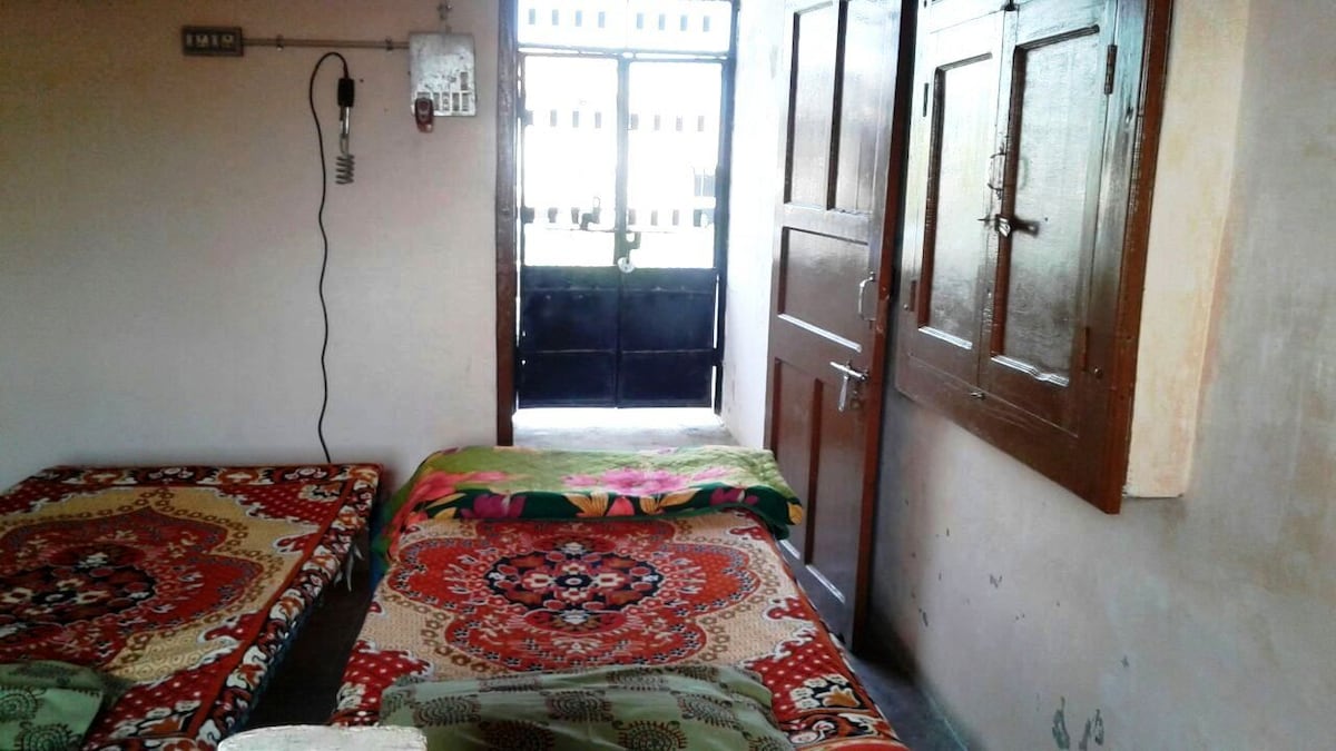 The room features two beds adorned with colorful bedspreads and decorative throws. A door leads to the outside, allowing natural light to enter. The walls are painted in neutral tones, and wooden cabinets provide storage options.